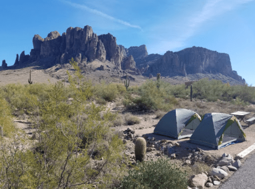 Two tents set up in a desert landscape with rocky mountains and cacti in the background under a clear blue sky.