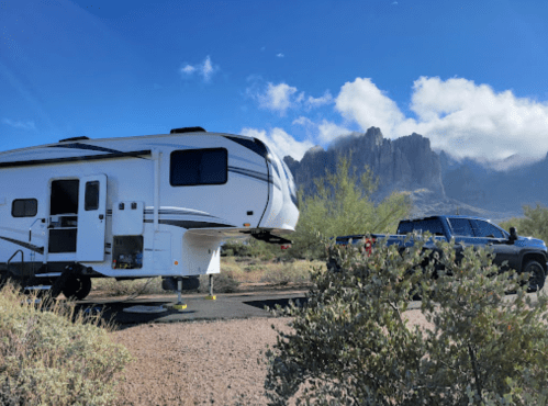 A white RV parked beside a pickup truck in a desert landscape with mountains and blue skies in the background.