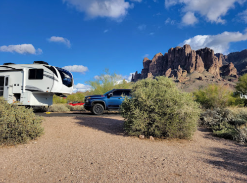 A camper and a blue truck parked near a rocky mountain landscape under a blue sky with scattered clouds.