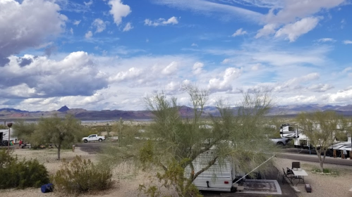 A desert landscape with sparse vegetation, RVs, and a cloudy blue sky in the background.