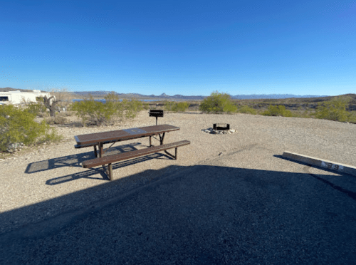 A picnic table and fire pit in a gravel area surrounded by sparse vegetation and mountains in the distance.