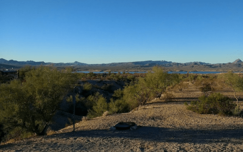 A serene desert landscape with sparse vegetation, mountains in the distance, and a calm body of water under a clear blue sky.