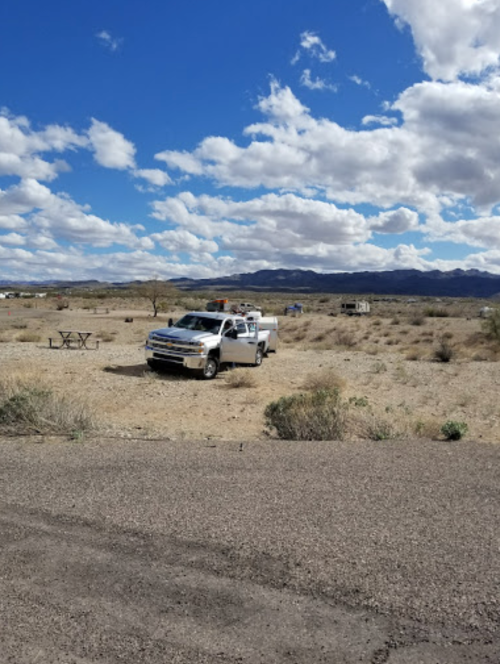 A pickup truck parked in a desert landscape with sparse vegetation and a picnic table under a cloudy sky.