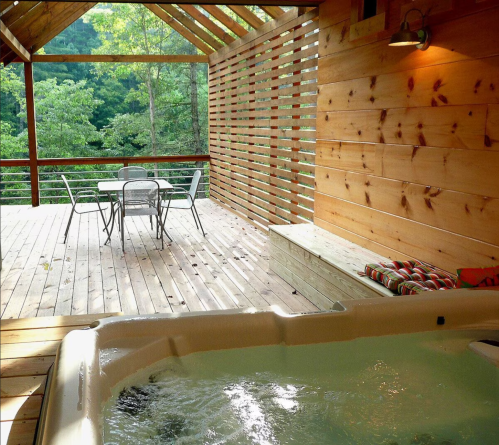 A wooden deck with a hot tub, table, and chairs, surrounded by greenery and natural light.