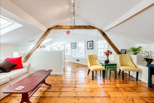 Bright, airy attic space with wooden floors, a sofa, two yellow chairs, and decorative plants.