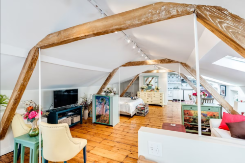 Bright, airy loft with wooden beams, a TV, cozy seating, and a bed, featuring a mix of modern and rustic decor.