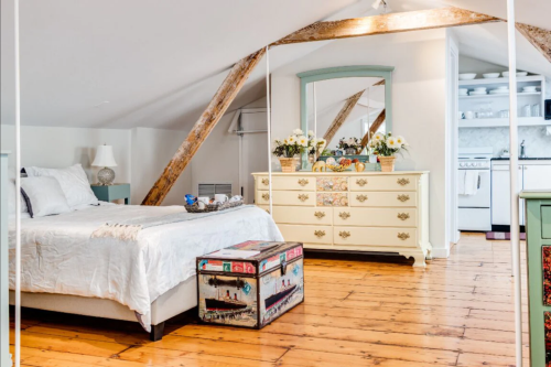Cozy attic bedroom with a bed, vintage dresser, and a kitchenette in the background, featuring wooden floors and beams.