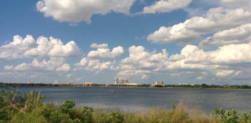 A serene lake scene with fluffy clouds above and a distant city skyline on the horizon. Greenery in the foreground.
