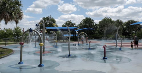 A splash pad with colorful water features, children playing, and palm trees under a partly cloudy sky.