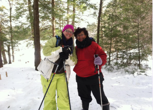 Two women smiling in winter attire, holding trekking poles, surrounded by snow and trees.