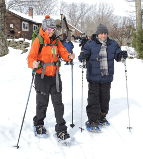 Two people snowshoeing in a snowy landscape, wearing winter gear and walking along a trail with trees in the background.