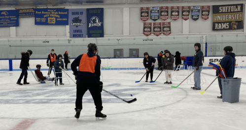 A group of people on an ice rink practicing hockey skills, with some wearing orange vests and others seated.