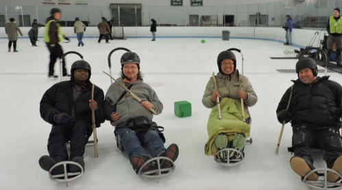 Four individuals sitting on sleds on an ice rink, smiling and holding sticks, with others skating in the background.