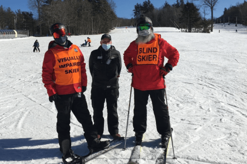 Two skiers wearing bright orange vests labeled "Visually Impaired Skier" and "Blind Skier" stand with an instructor on a snowy slope.