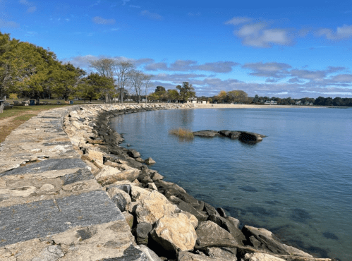 A serene waterfront scene with a rocky shoreline, calm water, and trees under a partly cloudy sky.