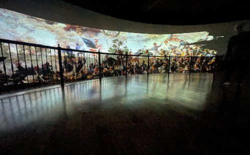 A panoramic mural depicting a historical battle scene, projected on a curved wall with a railing in the foreground.