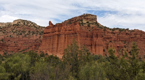 A rugged red rock formation rises above green vegetation under a cloudy sky.