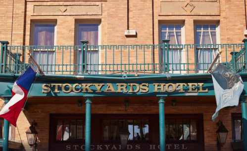 Exterior of Stockyards Hotel with flags and a brick facade, featuring a green balcony and large sign.