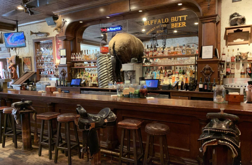 A rustic bar with wooden stools, a large buffalo head mounted above, and shelves filled with various bottles of alcohol.