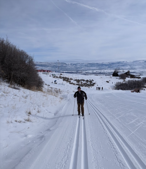 A person cross-country skiing on a snowy trail with mountains in the background and others skiing in the distance.