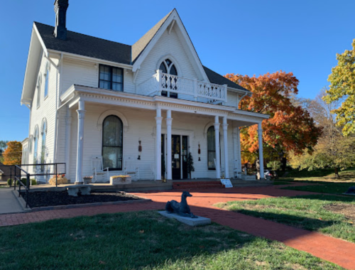 A white Victorian-style house with a porch, surrounded by colorful autumn trees and a grassy area.