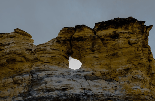 A rocky cliff with a large hole in the center, set against a cloudy sky.