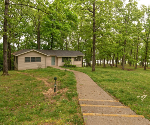 A single-story house surrounded by lush green trees, with a pathway leading to the entrance.