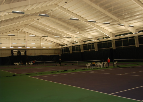 Indoor tennis court with multiple courts, chairs, and a high ceiling, illuminated by overhead lights.