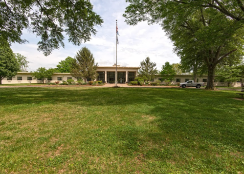 A grassy area in front of a building with a flagpole, surrounded by trees and a parked vehicle.