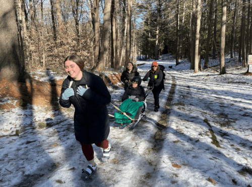 A group of four people enjoying a snowy forest trail, with one person being pulled on a sled.