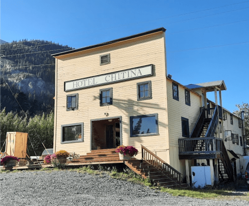 A rustic building labeled "Hotel Chitina" with a staircase, surrounded by greenery and mountains in the background.