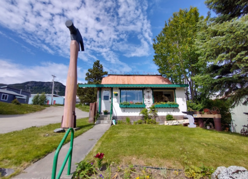 A quaint building with a hammer sculpture outside, surrounded by greenery and under a blue sky with clouds.