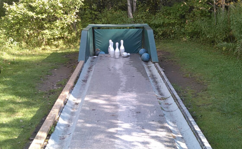 A bowling lane outdoors with three pins set up at the end and two blue bowling balls nearby.