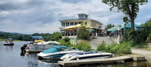 A lakeside restaurant with outdoor seating, surrounded by boats and greenery under a cloudy sky.