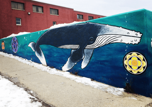 A colorful mural of a whale swimming in blue water, with decorative symbols on either side, against a snowy backdrop.