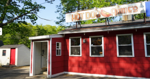 A red building with a sign reading "Hot Dog Annie's," featuring white windows and a small shed nearby.