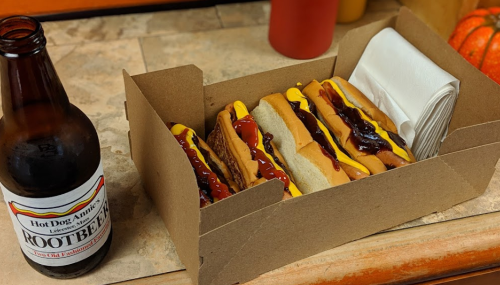 A takeout box with four hot dogs topped with mustard and ketchup, alongside a bottle of root beer.