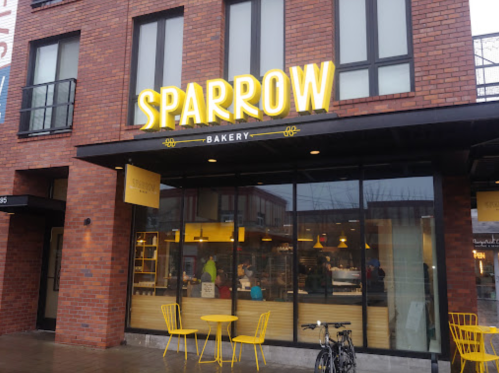 Exterior of Sparrow Bakery with a bright yellow sign, large windows, and yellow chairs outside.