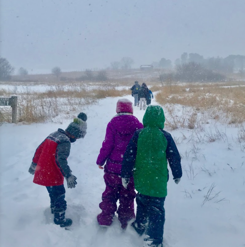Children in winter clothing walking on a snowy path, with snow falling and a distant group in the background.