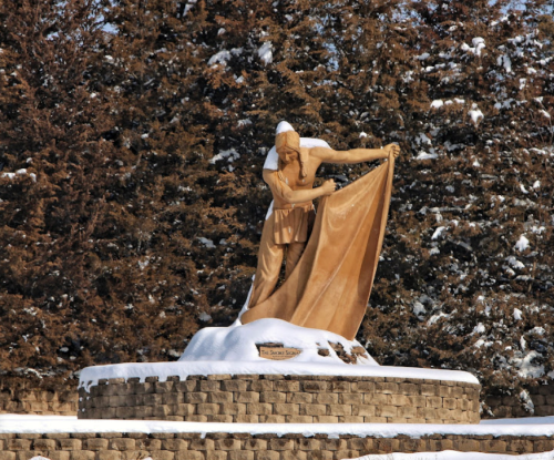 A snow-covered statue of a figure draping a cloth, surrounded by evergreen trees.