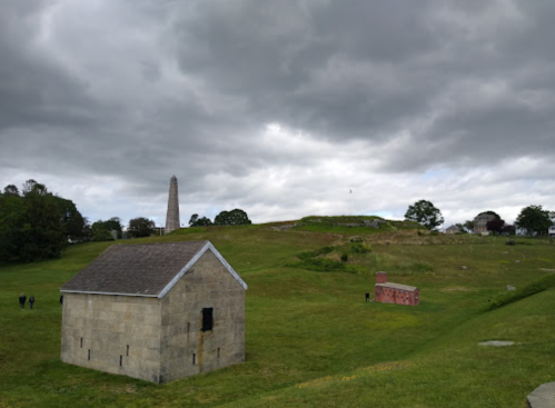 A grassy landscape with a stone building, a monument in the distance, and overcast skies.