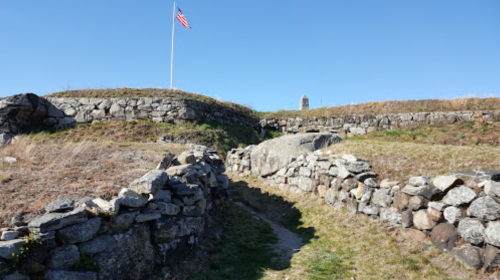Stone walls and pathways lead through grassy terrain, with an American flag flying in the background under a clear blue sky.