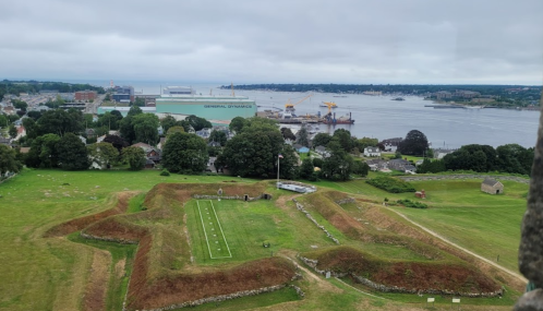 Aerial view of green fields with historical earthworks, a harbor, and industrial buildings in the background under a cloudy sky.