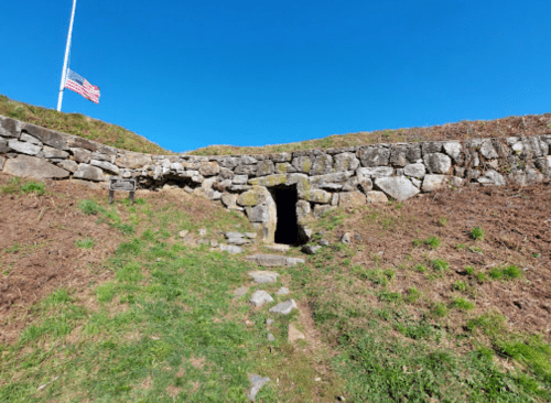 A stone entrance to a cave or tunnel, surrounded by grass, with an American flag flying in the background.