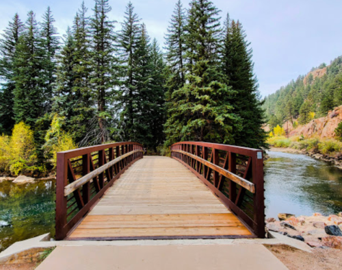 A wooden bridge spans a calm river, surrounded by tall evergreen trees and autumn foliage.