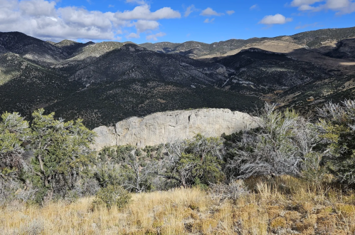 A panoramic view of rugged mountains and a rocky cliff under a blue sky with scattered clouds.