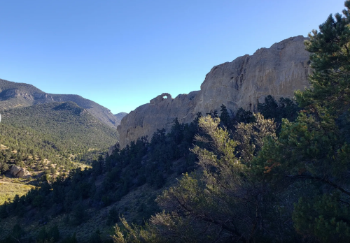A rocky mountain landscape with a distinctive arch formation, surrounded by trees and rolling hills under a clear blue sky.