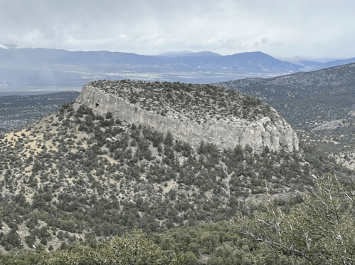 A rocky outcrop rises above a valley, surrounded by sparse vegetation and distant mountains under a cloudy sky.
