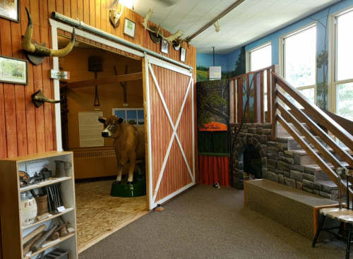 A rustic museum interior featuring a cow model, wooden decor, and a staircase leading to a nature-themed area.