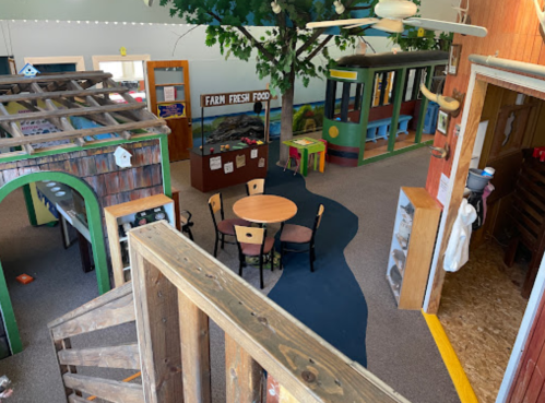 A colorful indoor play area with a tree, play structures, and a central table surrounded by chairs.